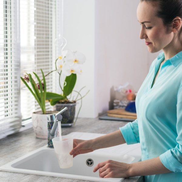 Portrait of a young woman gaining a glass of clean tap water in the kitchen in front of the window.